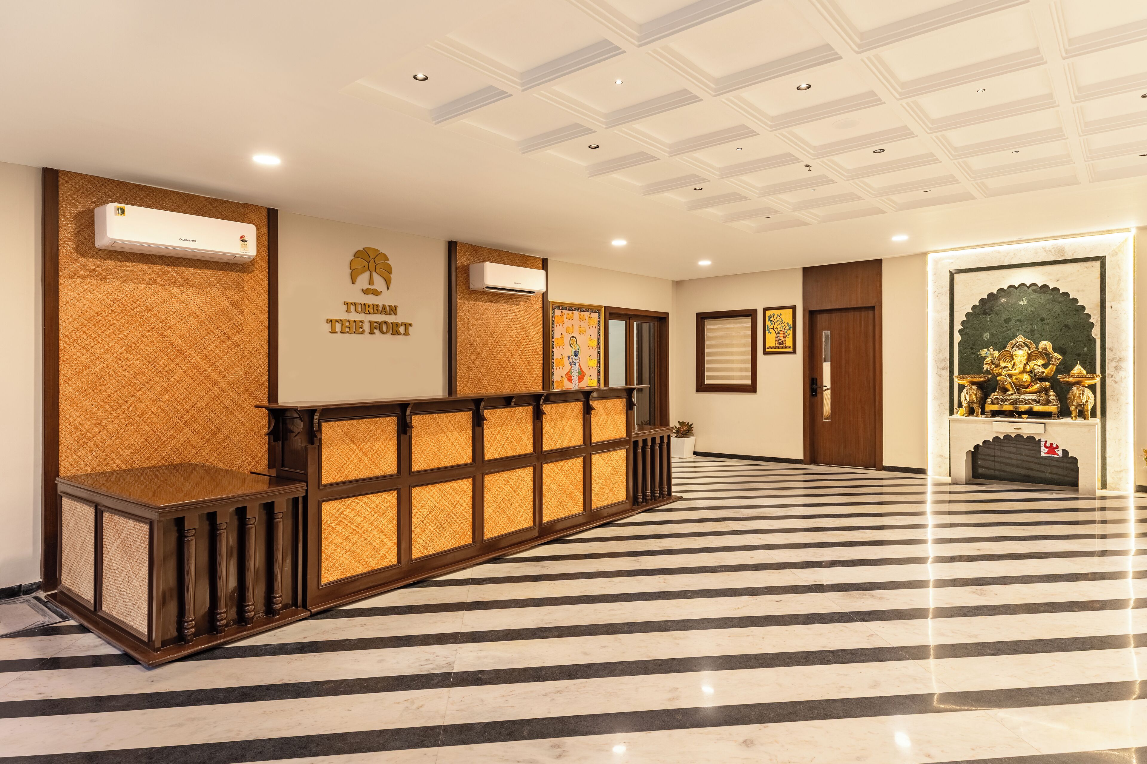A hotel reception area with a check-in counter, air conditioning units, and a decorative wall with a logo.
