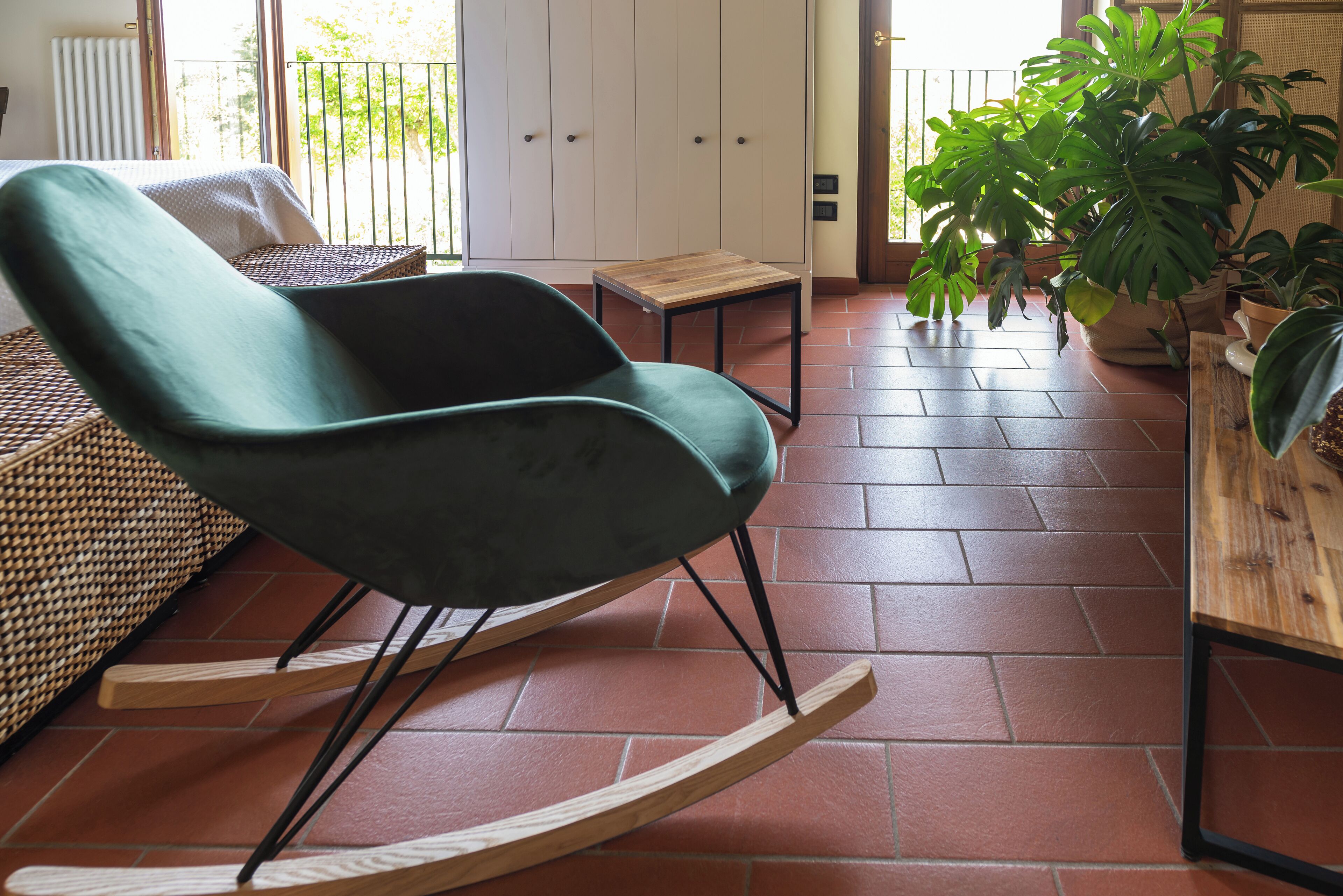 Un fauteuil à bascule, une petite table et des plantes en pot dans une pièce dont le sol est recouvert de carreaux, avec une vue sur l’extérieur par une porte.