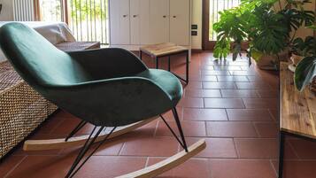 Un fauteuil à bascule, une petite table et des plantes en pot dans une pièce dont le sol est recouvert de carreaux, avec une vue sur l’extérieur par une porte.