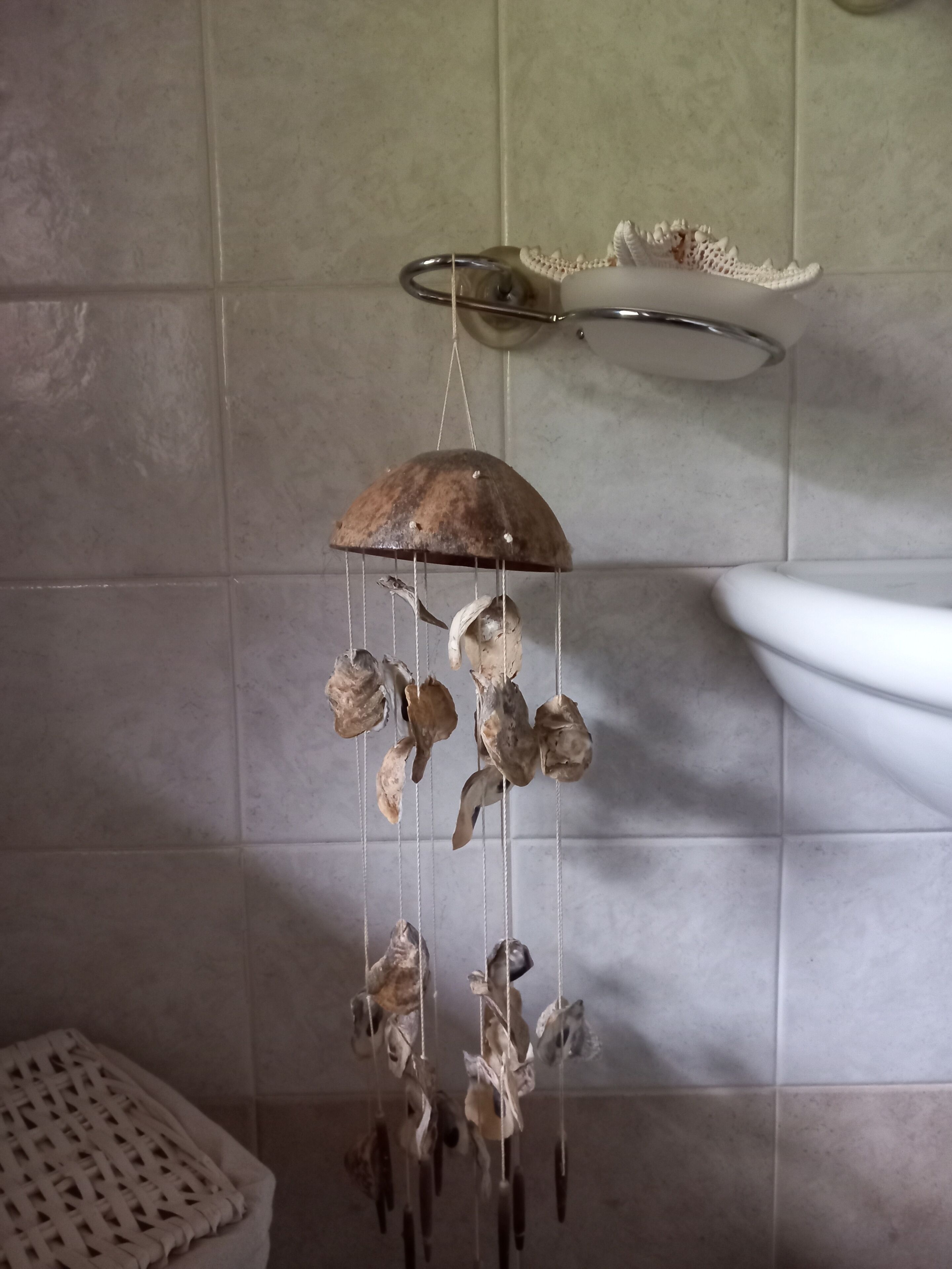 Ein hängendes Windspiel mit getrockneten Blättern und einer Metallschüssel, befestigt an einem Haken in der Decke eines Badezimmers mit gefliesten Wänden und einem weißen Waschbecken.