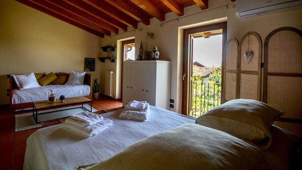 A hotel room with a wooden ceiling, a bed with white linens, a sofa with cushions, a small table with a vase, and a view through an open door.