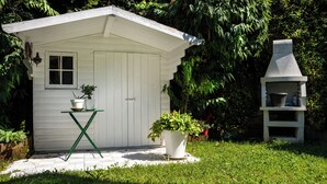 A small white shed with a window and a door, a green folding table with a potted plant, and a stone barbecue grill.
