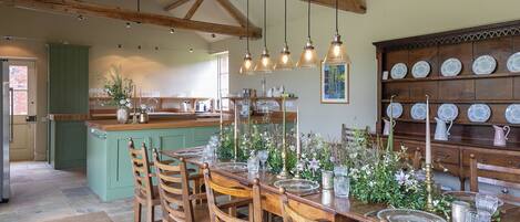 A dining area with a wooden table set for six, surrounded by chairs. A kitchen with a green island and wooden beams is visible in the background.