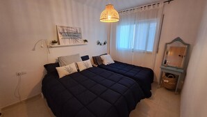 Ett sovrum med två sängar, en hängande lampa, en vägghylla med dekorativa föremål och ett fönster med tunna, genomskinliga gardiner.