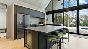 A modern kitchen with a large island, bar stools, and a view of a snowy landscape through large windows. - Luxury Home | Ski In / Ski Out | 5 Bed / 4 Bath (Whitefish)