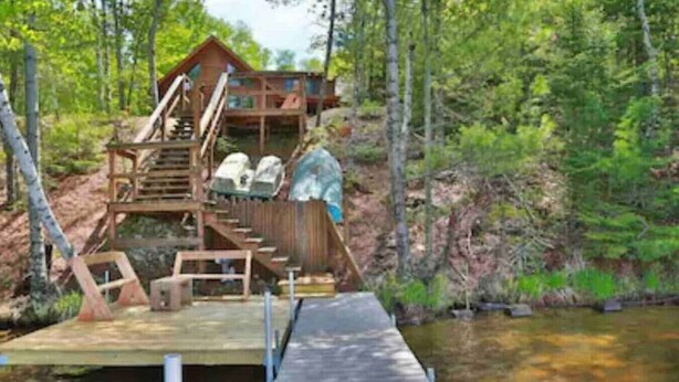 Une cabane en bois avec un escalier menant à une terrasse surplombant une étendue d’eau, le tout entouré d’arbres.