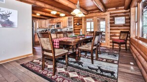 L’intérieur d’une cabane en rondins comprend un coin repas avec une table et des chaises en bois, une cuisine avec des meubles en bois et une grande fenêtre qui laisse entrer la lumière naturelle.