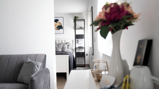 Una sala de estar moderna con un sofá gris, una mesa de centro blanca y un jarrón con flores. Hay un escritorio con una computadora y una estantería con plantas.