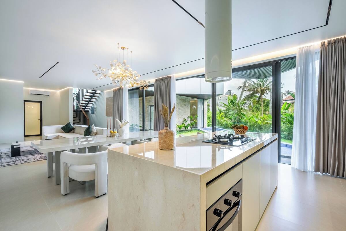 A modern kitchen with a central island, large windows, and a view of greenery outside.