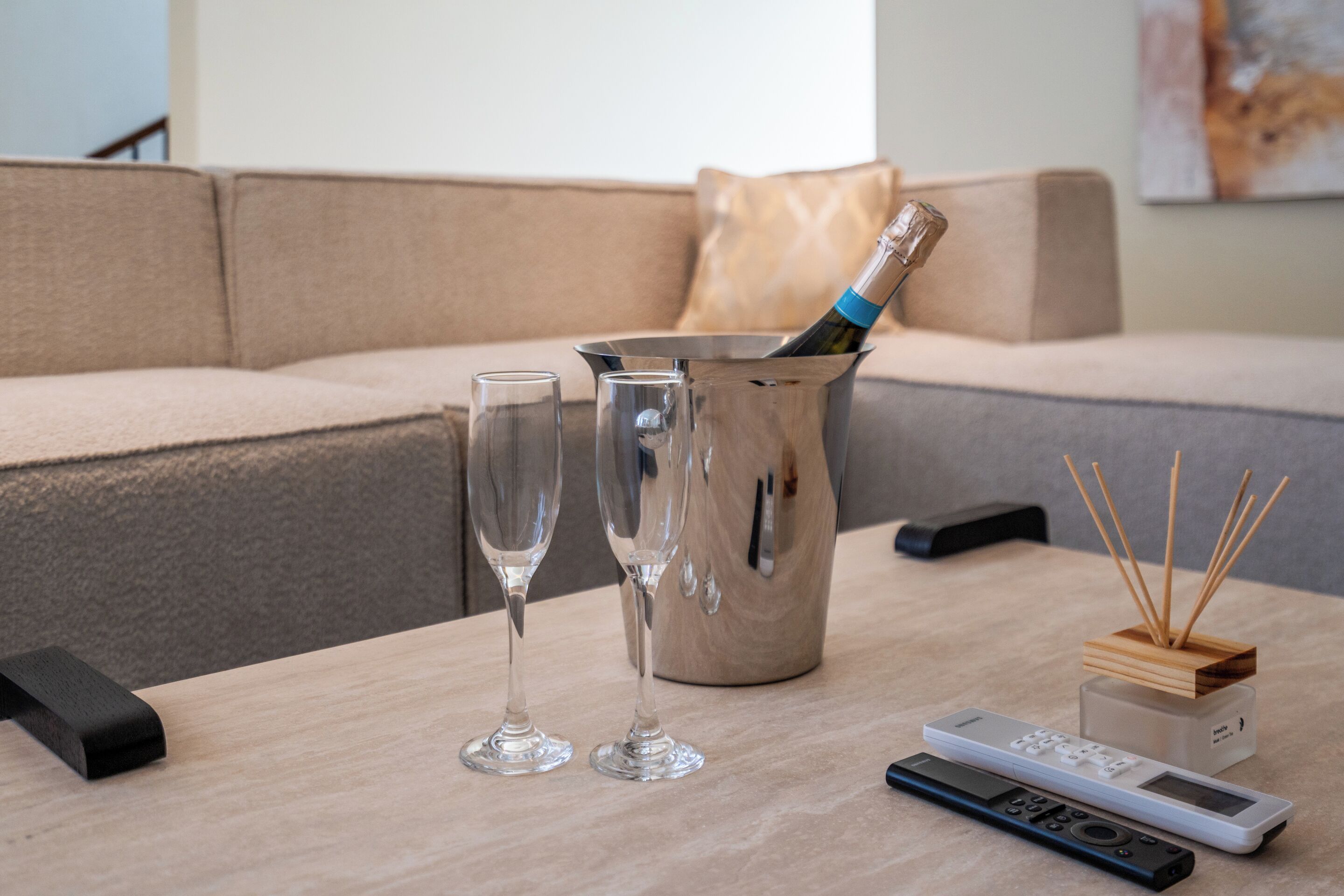 Ein modernes Wohnzimmer mit einem beigen Sofa, einem Holztisch, zwei Sektgläsern, einer Champagnerflasche, einer Fernbedienung und einem Reed-Diffusor.
