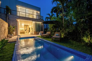 Una casa moderna de dos pisos amb una piscina, mobiliari d'exterior i vegetació abundant.