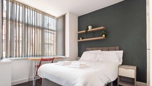 Quarto de hotel moderno com uma cama grande, uma escrivaninha e uma cadeira. Há duas prateleiras com plantas na parede.