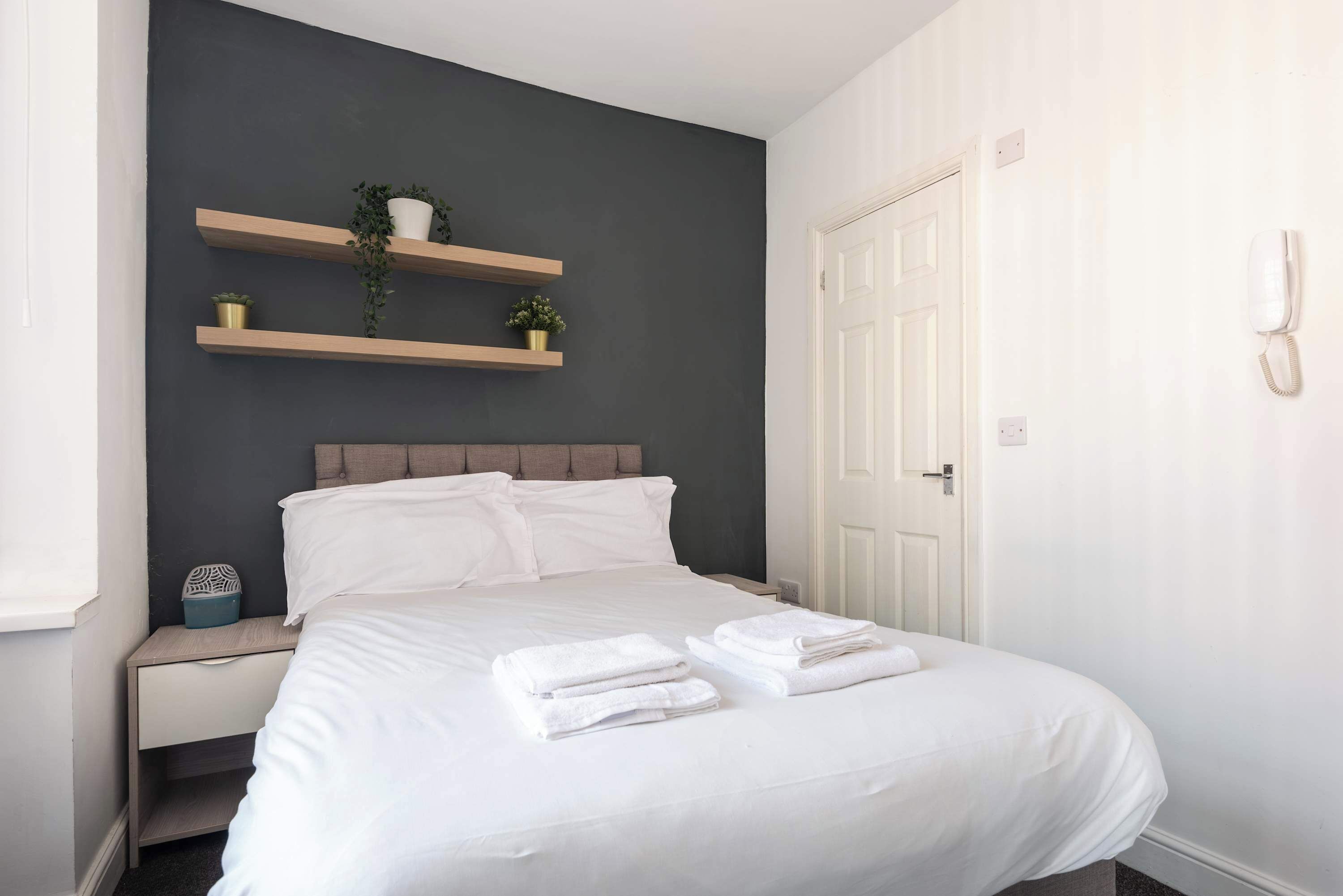 A neatly made bed with white linens and towels, a bedside table with a decorative item, and a wall-mounted shelf with plants.
