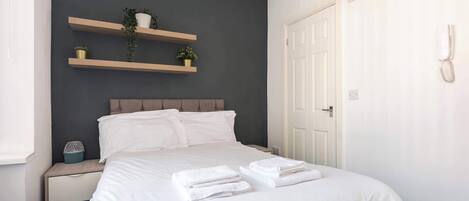 A neatly made bed with white linens and towels, a bedside table with a decorative item, and a wall-mounted shelf with plants.