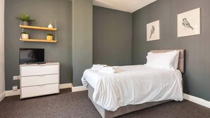 A modern bedroom with a bed, a TV on a stand, a dresser, and wall-mounted shelves with plants.
