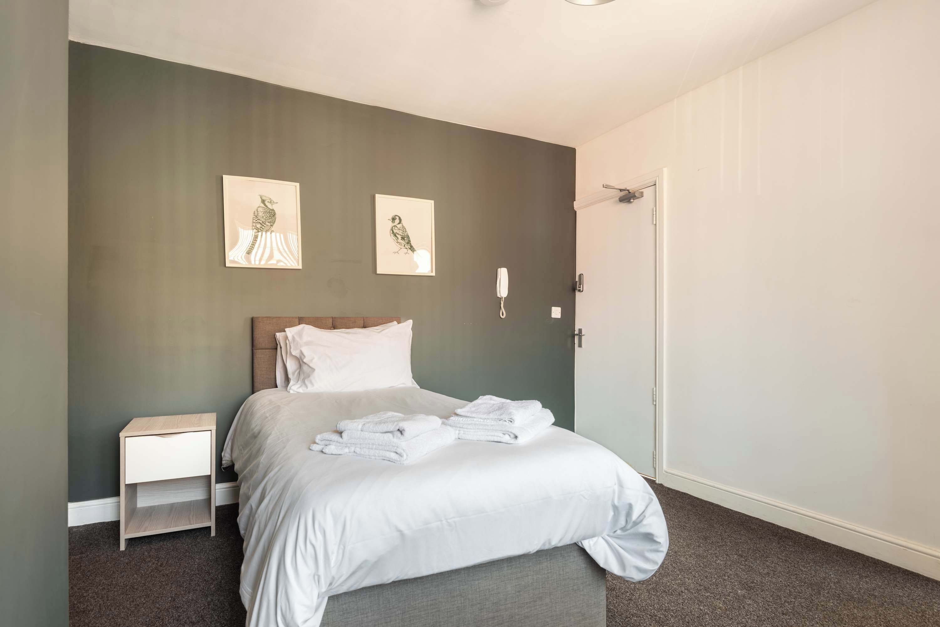 Ein Schlafzimmer mit einem Bett, einem Nachttisch, zwei an der Wand befestigten Lichtschaltern und zwei gerahmten Bildern.