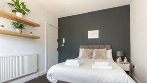 A hotel room with a bed, bedside table, lamp, and wall-mounted shelf with plants. - Convenient Budget Room in Shared House w Kitchen (Doncaster)