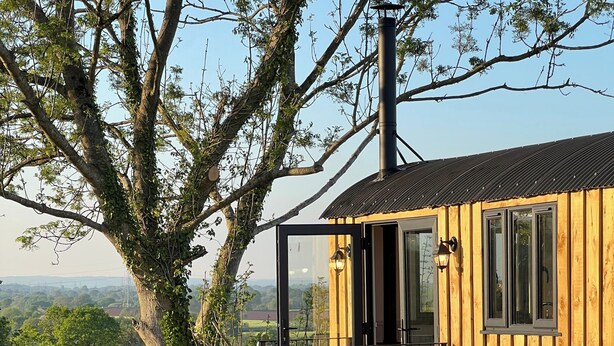 A wooden cabin with a glass door, a hot tub, and a patio with metal chairs. - Whimple Shepherd Hut, Sleeps 2, Wood Fire & Garden (Exeter)