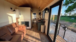 A cozy room with a wooden ceiling, a sofa, a radiator, and a view of a garden through a glass door. - Whimple Shepherd Hut, Sleeps 2, Wood Fire & Garden (Exeter)
