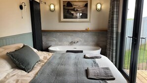 A bedroom with a large bed, a bathtub, and a view of the outdoors. - Whimple Shepherd Hut, Sleeps 2, Wood Fire & Garden (Exeter)