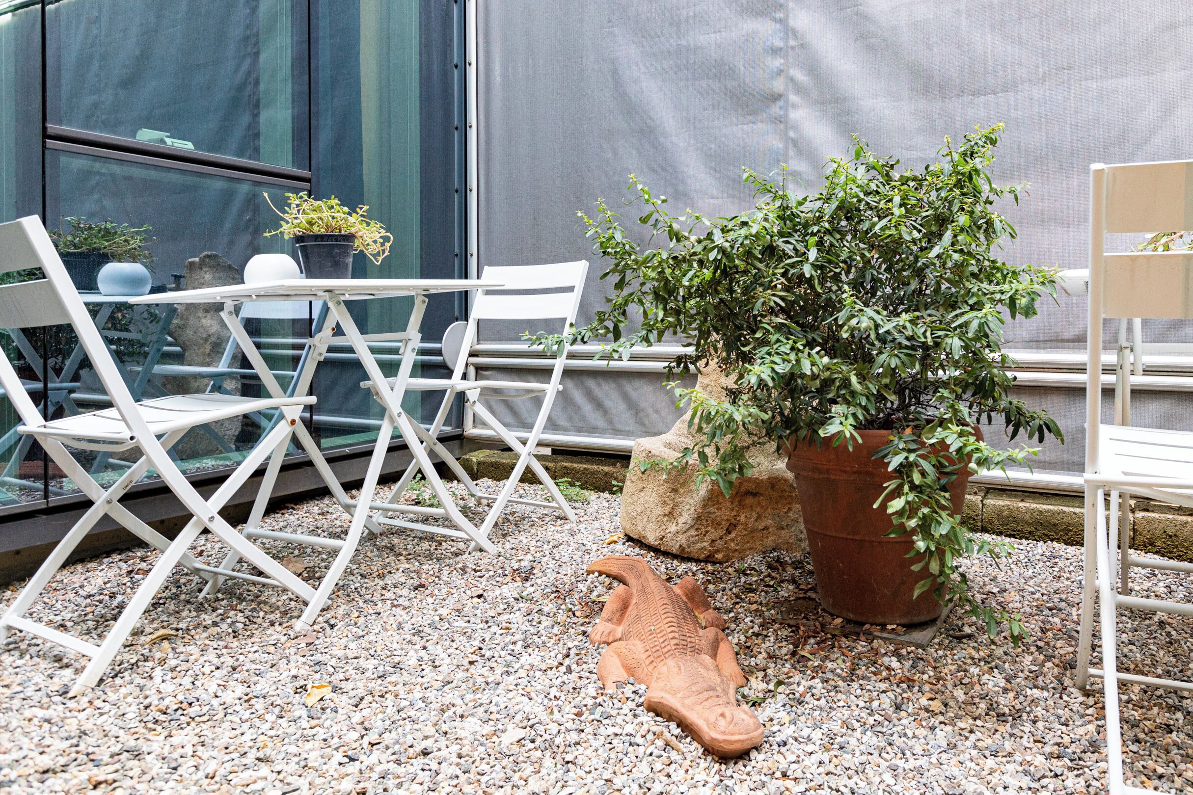 A seating area with white chairs and a table, a large potted plant, and a decorative fish sculpture on a gravel surface.