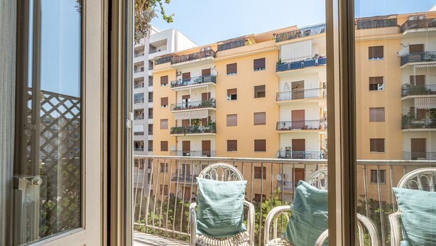 Un balcon agrémenté de fauteuils et de coussins en osier, d’où l’on peut admirer une rangée d’immeubles à plusieurs étages, chacun doté d’un balcon.