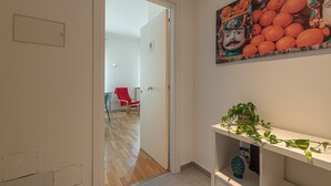 Un couloir avec un plancher en bois, une chaise rouge, une porte blanche et une étagère fixée au mur sur laquelle se trouve une plante.