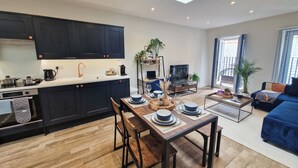 A modern kitchen with dark cabinets, a dining area with a wooden table, and a living room with a blue sofa and a TV. - Bright 1-Bed Flat Near Fulham Broadway Station (London)