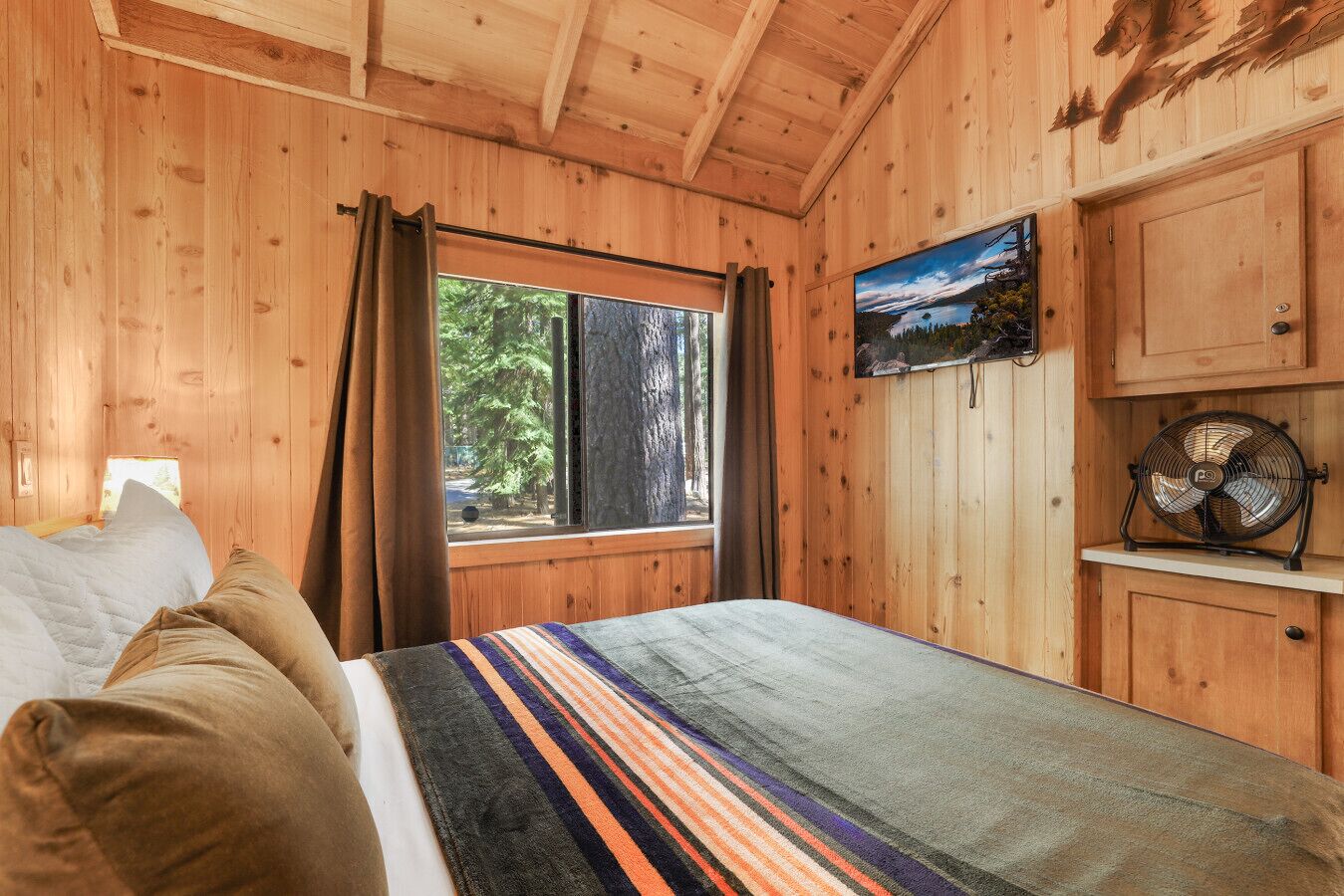 Een houten kamer met een bed, een raam met gordijnen, een schilderij en een ventilator.