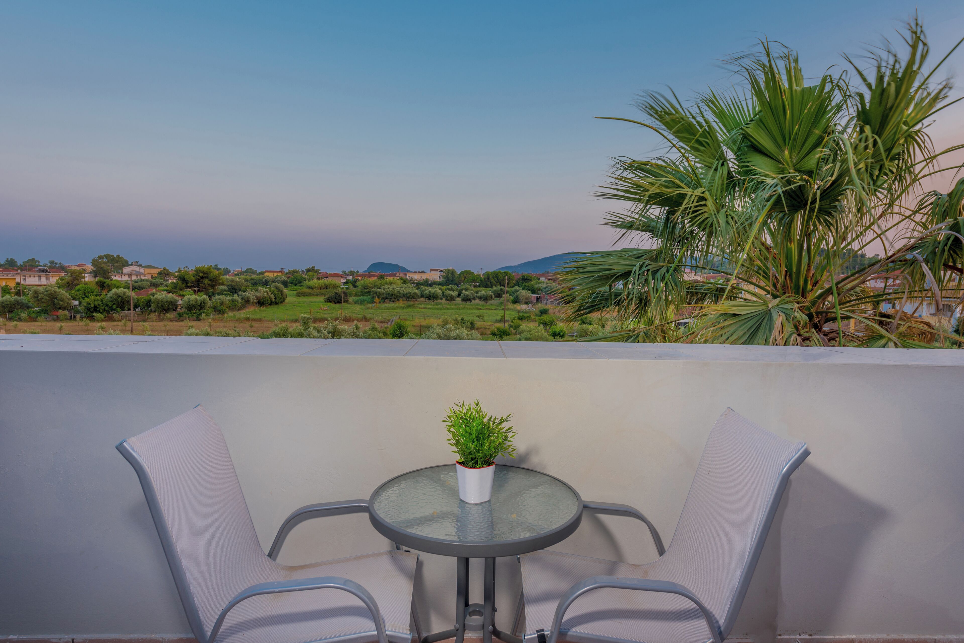 A balcony with a glass table and two chairs, overlooking a coastal landscape with houses and greenery.
