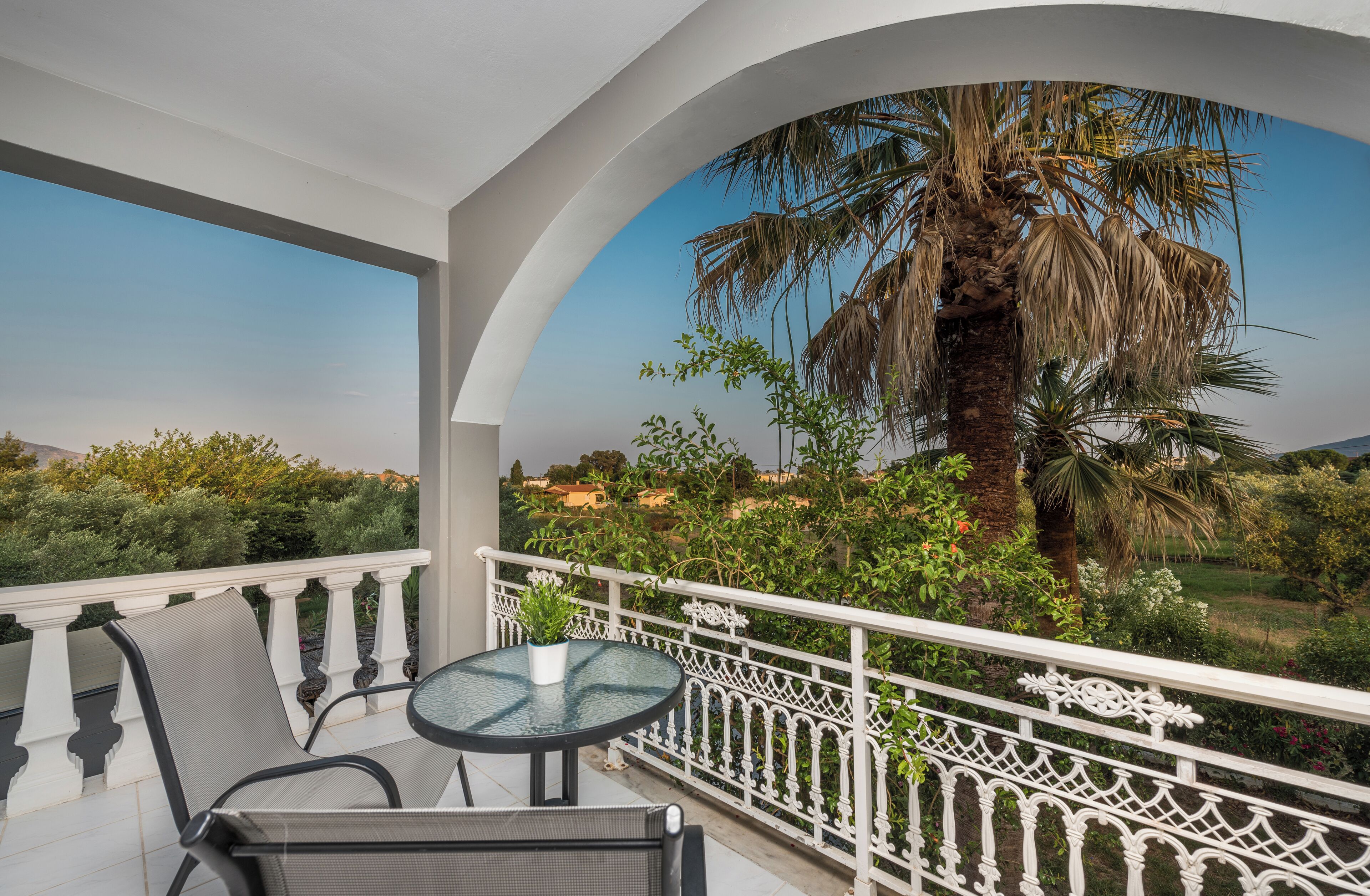 A balcony with a round table and chairs, white balustrade, and a view of a palm tree and greenery.