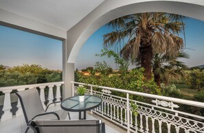 Un balcón con una mesa redonda y sillas, barandilla blanca, y vistas a una palmera y follaje.