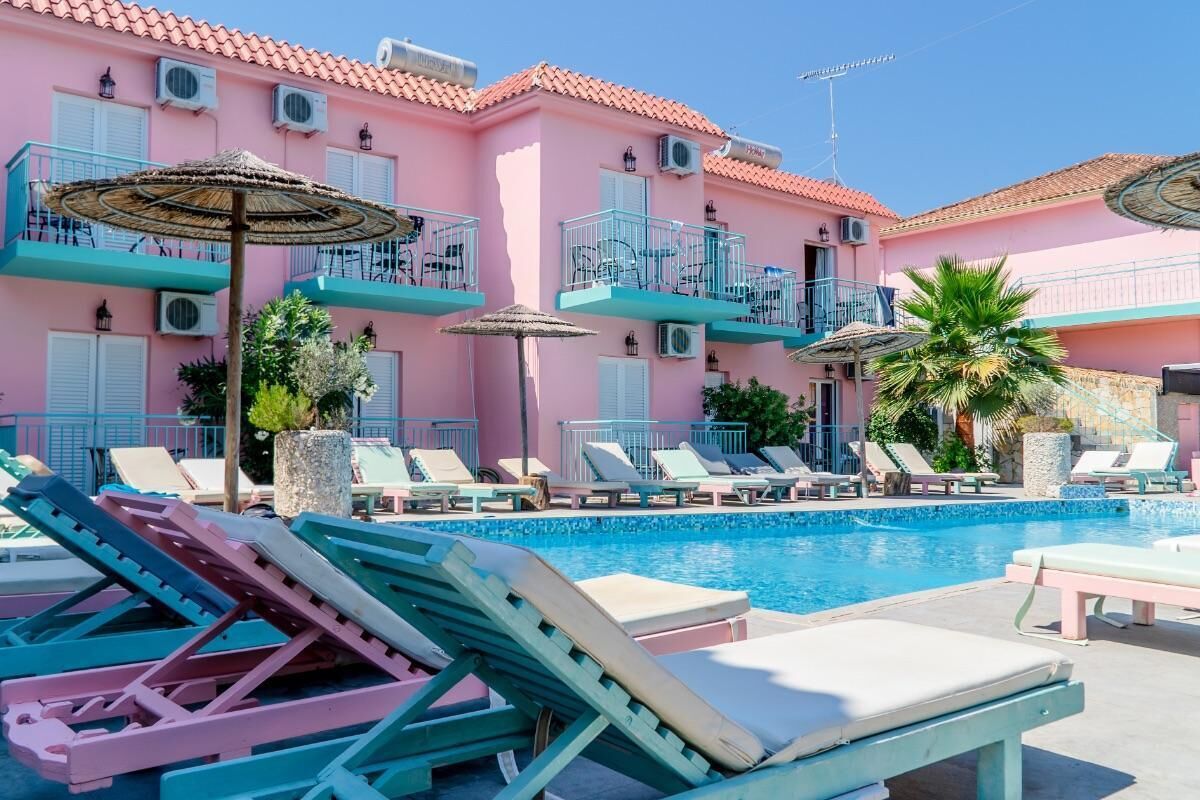 A pink building with a pool, lounge chairs, and umbrellas.