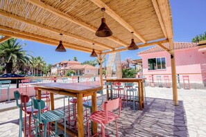 An outdoor seating area with wooden tables and chairs, a bamboo roof, and a view of buildings in the background. - MAUI HOTEL LAGANAS (Laganas)