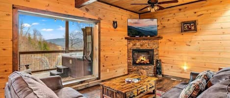 A cozy cabin interior with a fireplace, leather sofas, a wooden coffee table, and a large window overlooking a scenic landscape.