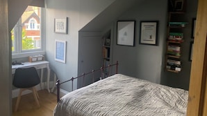 A bedroom with a bed, a desk, and a chair. There are framed pictures on the wall and a bookshelf with books.
