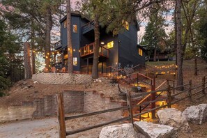 A modern house with a stone staircase, wooden deck, and string lights. - EV-Charged Treetop Cabin w/ Game Room & Decks (Lake Arrowhead)