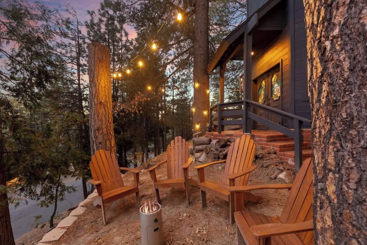 Un patio in legno con poltrone Adirondack, un braciere e luci decorative, sullo sfondo alberi e un edificio.