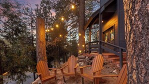 A wooden deck with Adirondack chairs, a fire pit, and string lights against a backdrop of trees and a building.