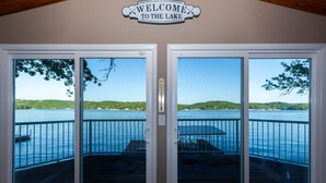 A view through a set of glass doors with a 'Welcome to the Lake' sign above, leading to a deck with a railing overlooking a body of water and greenery. - Newly Listed! Lakefront Home Private Dock with Huge Views (Camdenton)