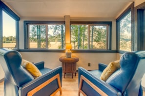 A room with two blue leather chairs, a small wooden table with a lamp, and large windows with a view of trees.