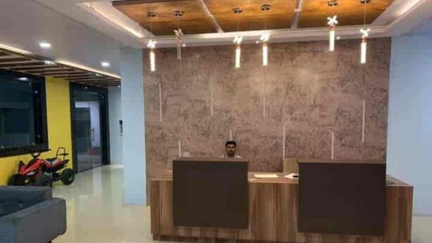 A reception desk with two computer monitors, a wooden ceiling with recessed lighting, and a grey sofa in the foreground. - Hotel Crown Resort (Satna)