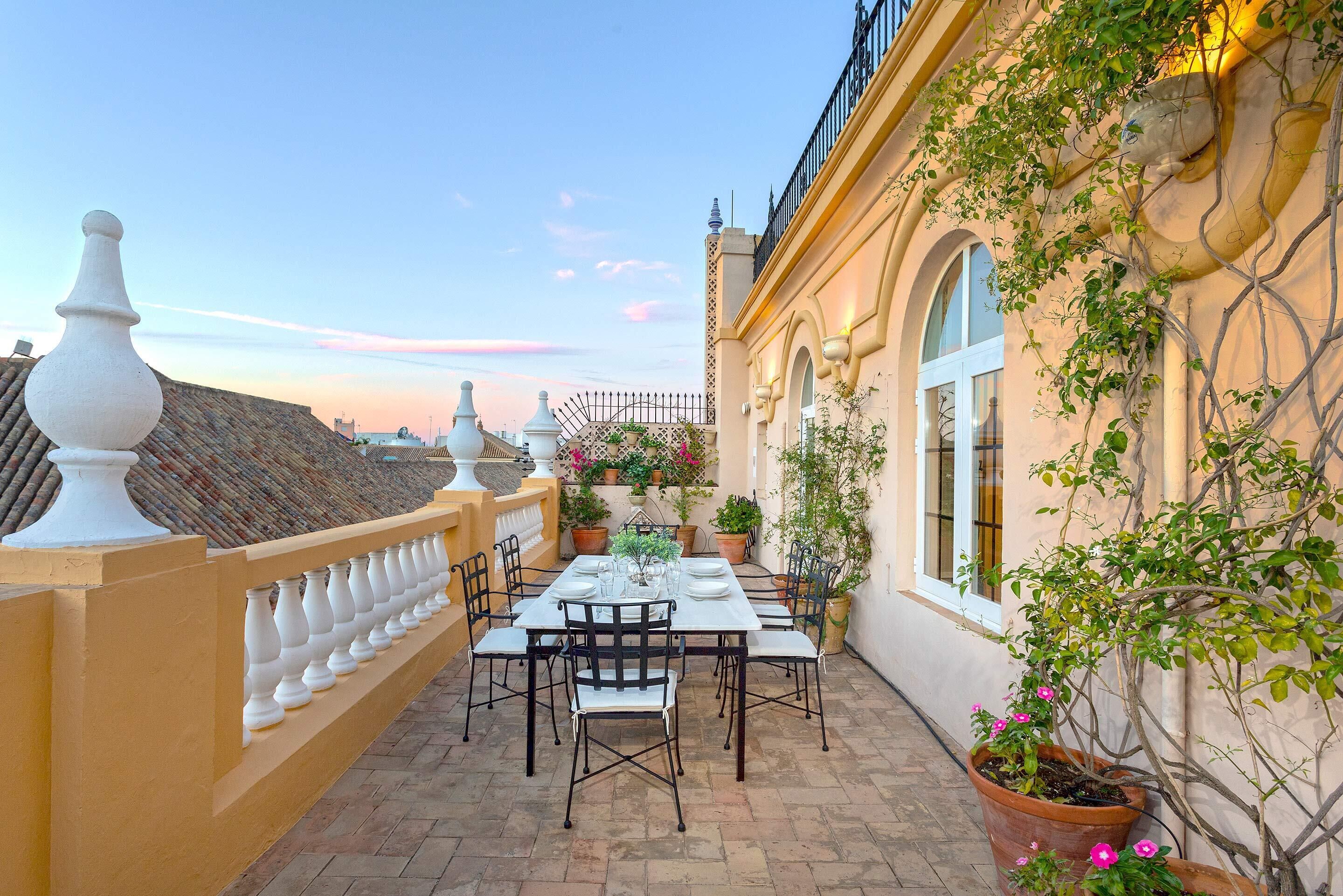 A rooftop terrace with a dining table set for six, surrounded by chairs, potted plants, and a decorative railing.