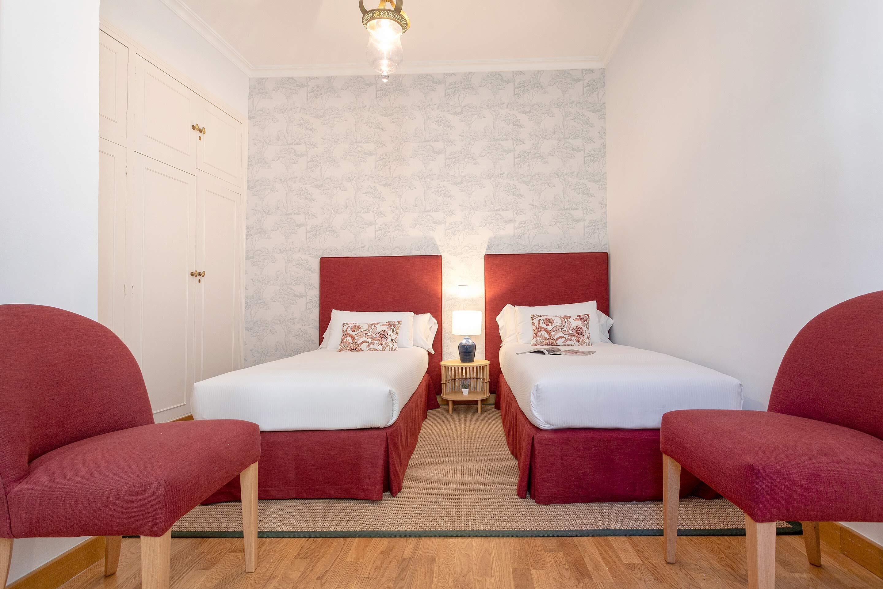 A room with two single beds, a small table with a lamp, and two red chairs.