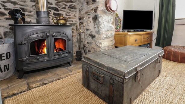 A stone fireplace with a black stove, a wooden chest, and a television on a wooden cabinet. - Cobweb Cottage (Caernarfon)