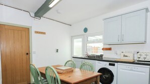A compact kitchen with a wooden table, green chairs, a washing machine, and white cabinets. - Cobweb Cottage (Caernarfon)