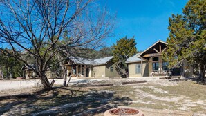 A rustic lodge with a stone fire pit, wooden deck, and a large open yard with trees.