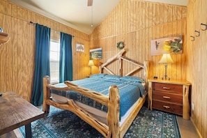 A wooden-paneled room with a bed, a wooden chair, a small table, and a nightstand with a lamp.