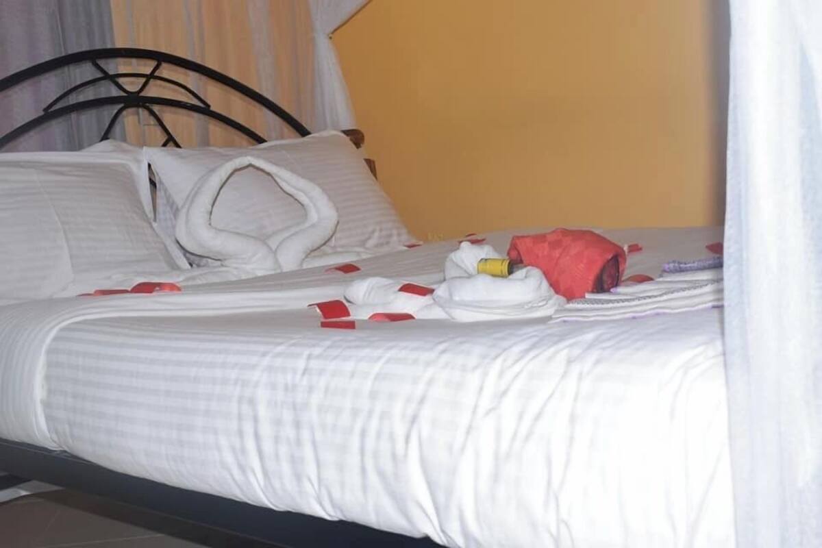 Cama bem arrumada com lençóis brancos e detalhes vermelhos, cabeceira de metal preto e parede amarela lisa ao fundo.