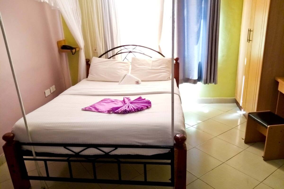 Uma cama de solteiro com roupa de cama branca e toalha roxa, cabeceira de madeira e uma janela com cortinas brancas.